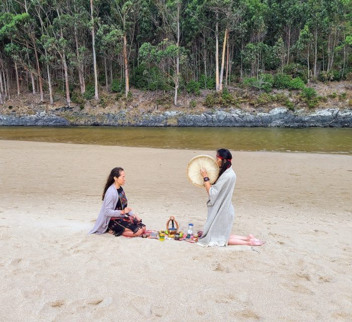 Two women engaged in a shamanic healing ritual on a sandy beach, with one playing a drum and the other sitting cross-legged surrounded by ritual items, against a backdrop of trees and a river.