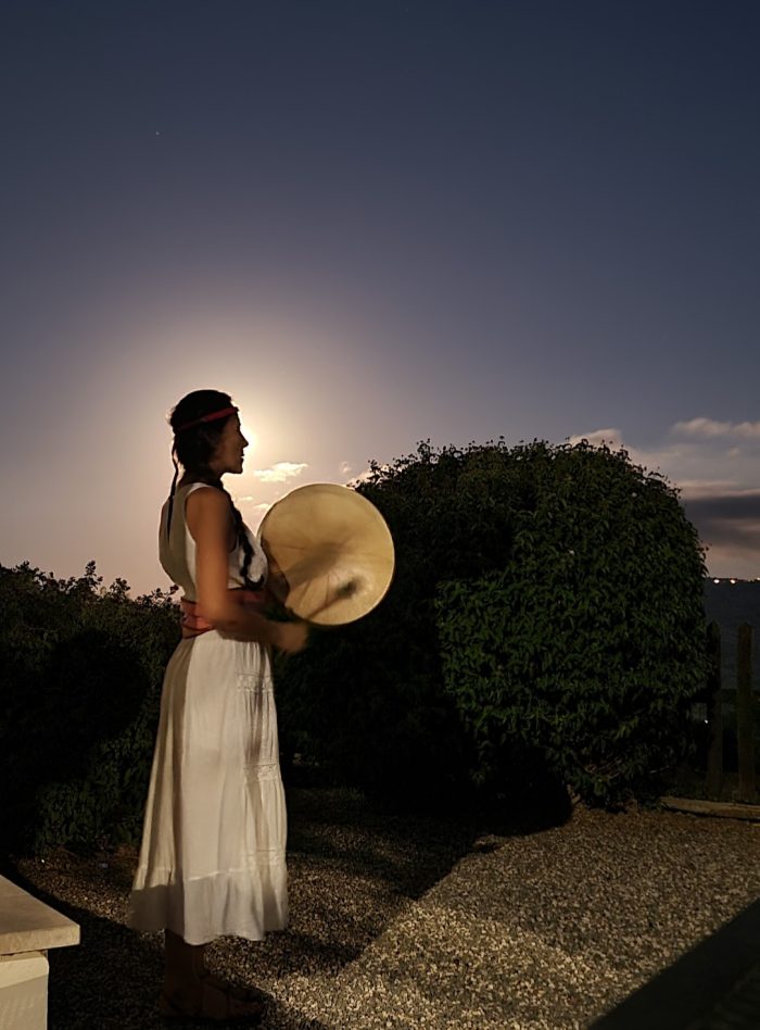 A woman playing a drum during a Full Moon shamanic ritual, silhouetted against a full moon with a background of trees.