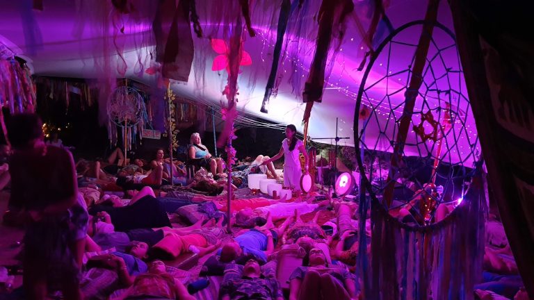 Carolina of Luminous Five leads a sound healing ceremony under a vibrant canopy bathed in pink and purple light, surrounded by dreamcatchers and colorful fabrics. Participants lie in a deep meditative state, immersed in the soothing sounds of drums and singing bowls, creating a sacred and transformative space filled with spiritual energy.