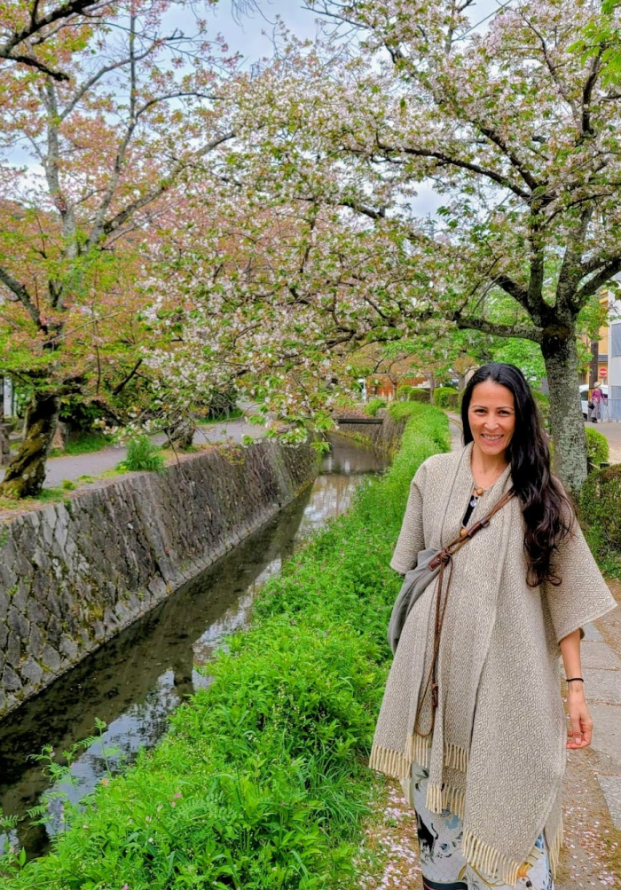 Carolina of Luminous Five stands beside a serene canal lined with blooming cherry blossom trees, wearing a light gray long poncho and smiling, radiating joy and connection with nature.